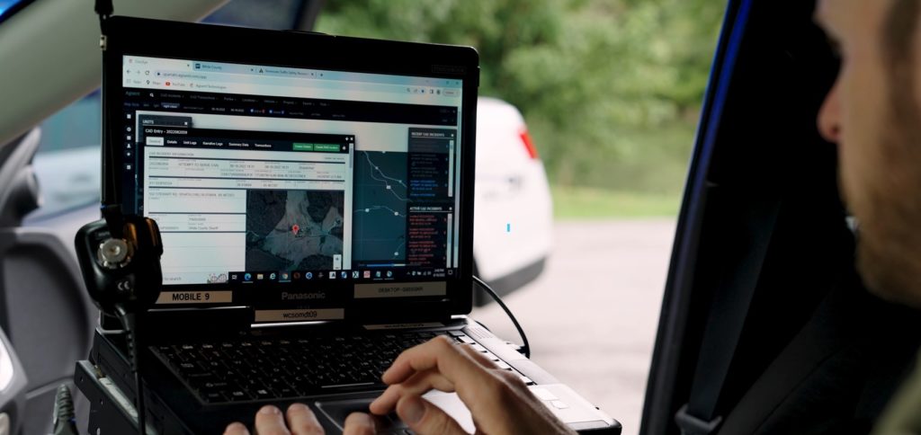 Image of an officer sitting in a patrol car. The image is focused on the laptop computer screen, where we can see the officer is using an RMS on-the-go.