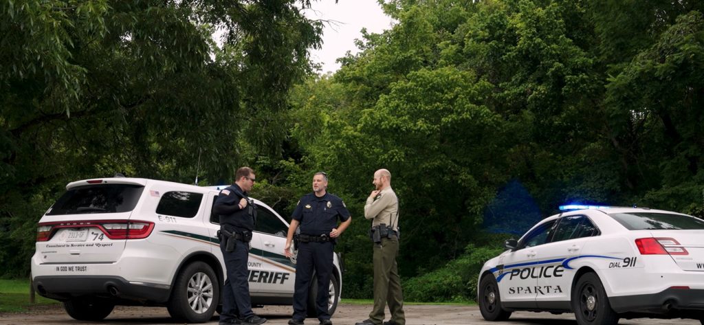 Three law enforcement officers standing talking with one another, between two patrol cars. They are outside, surrounded by trees.