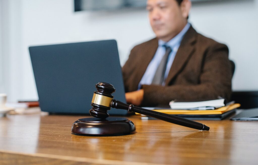An attorney/prosecutor/courtroom official uses digital tools for evidence documentation. In this image, a man sits behind a laptop working. In front of the laptop, on the man's desk, sits a gavel, representative of the justice system.