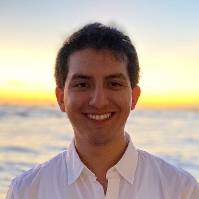 A headshot of Hector Nevarez. he stands outside smiling, with a sunset and the sea in the background.