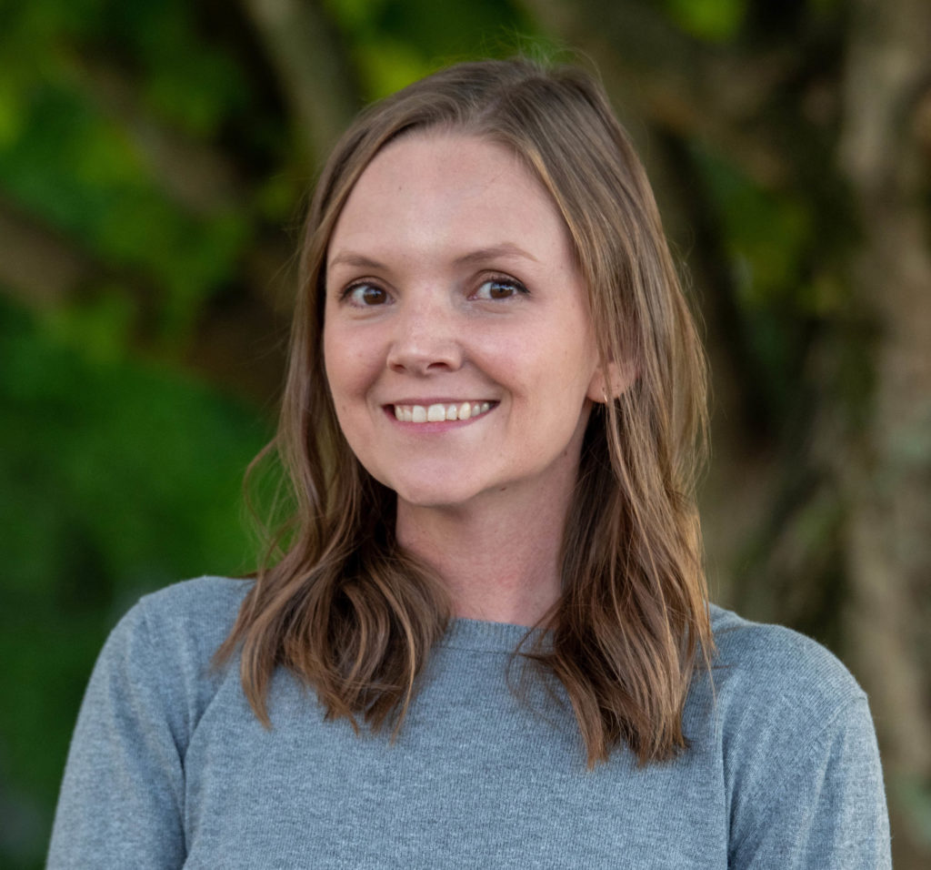 A smiling headshot of Christie Pearce. The photo is from shoulders up and is shot outdoors in front of some trees.