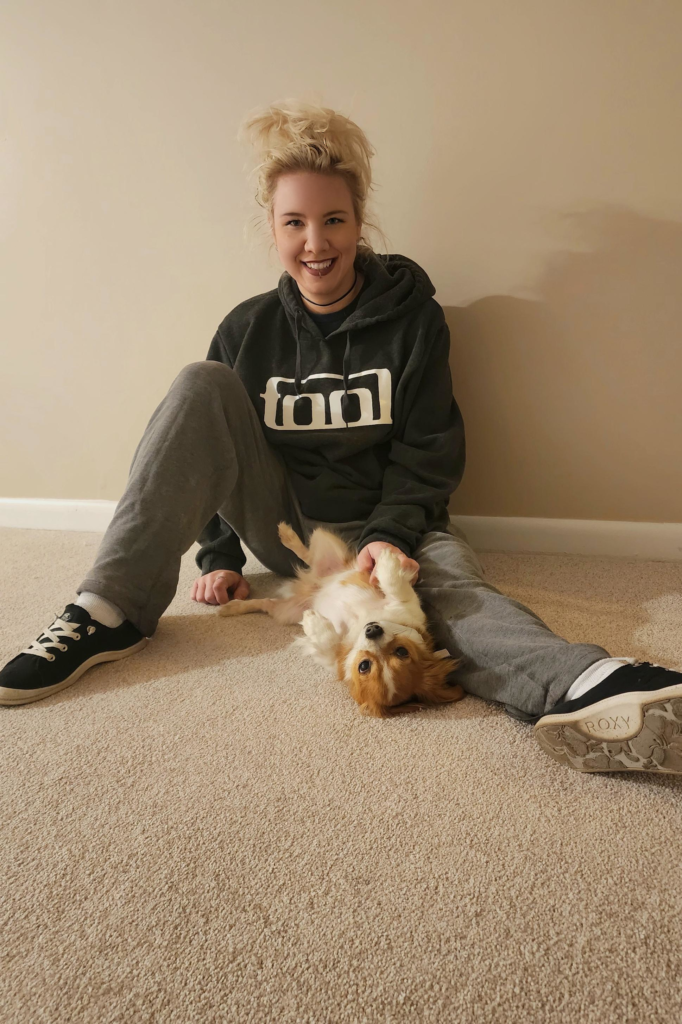 A photo of Martina Gasser. She sits smiling on the floor, with her dog laying on its back in front of her.