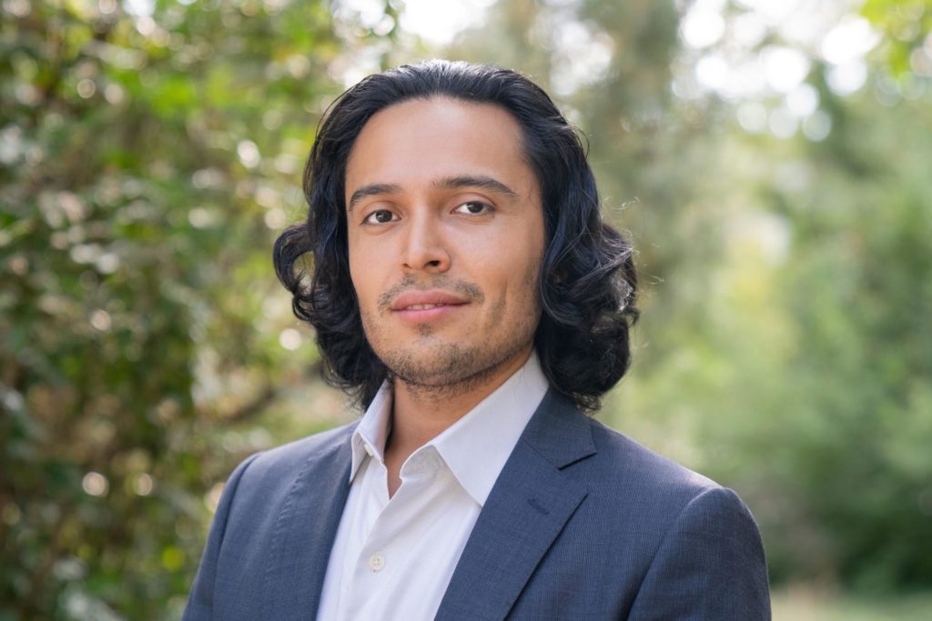 A headshot of Alfredo Velasco. He is wearing a suit and standing outside with trees in the background.